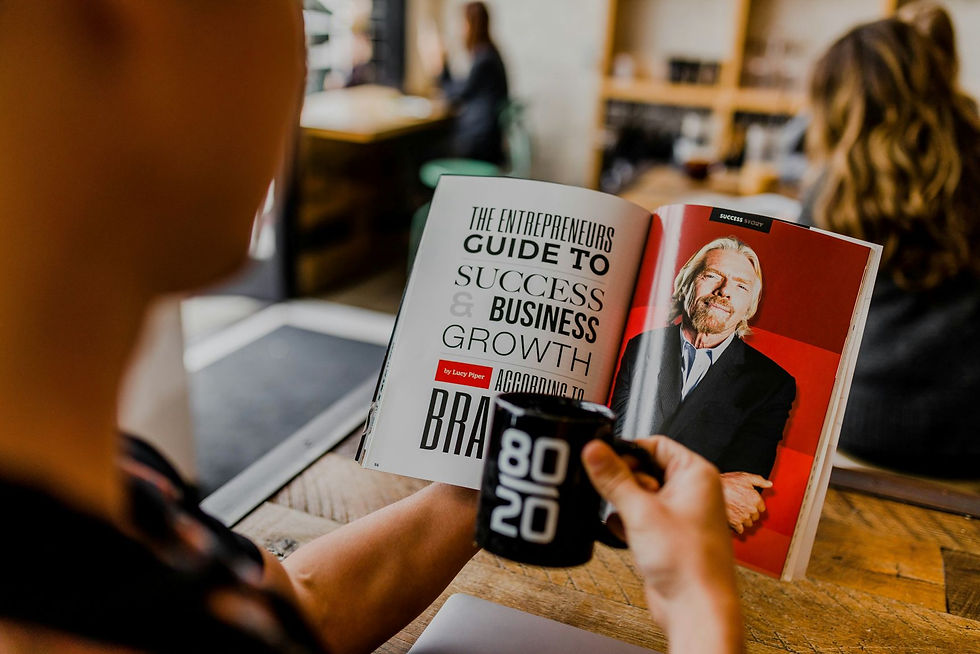 Person reading a business magazine, holding a black mug labeled "80 20." Magazine text: "The Entrepreneur's Guide to Success." Cozy cafe setting.