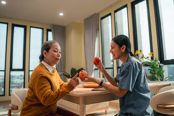 Two women chat at a table in a bright room, smiling while holding apples, with large windows and plants in the background.