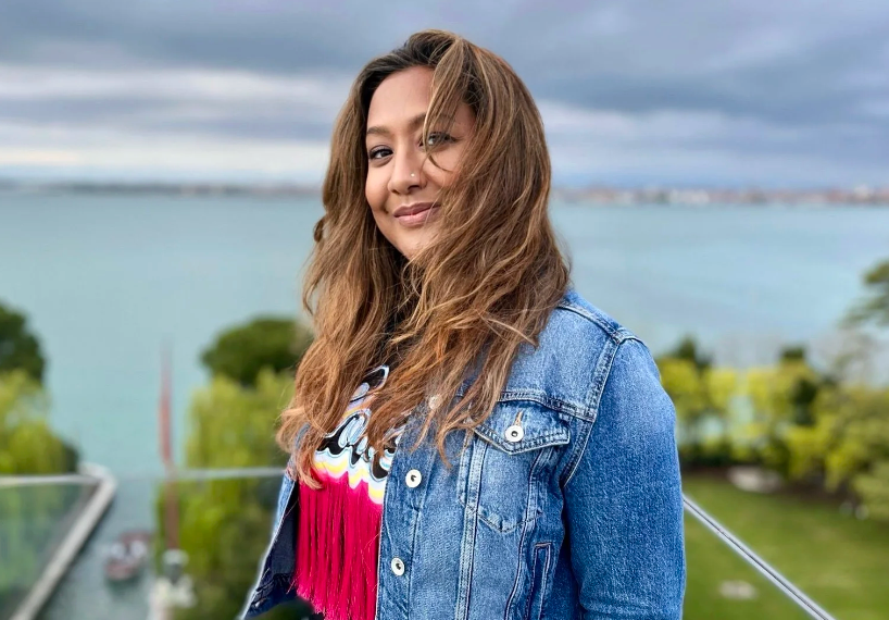 Woman in denim jacket smiles by a lake. Overcast sky and greenery in background. She's wearing a colorful top. Mood is relaxed.