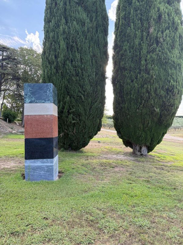 Striped stone pillar stands in grass between two tall cypress trees under a bright sky.