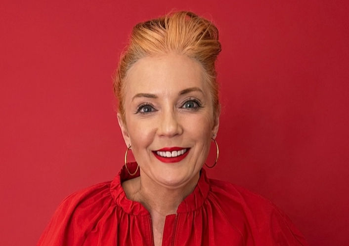 Smiling woman with red lipstick and top against a matching red background, wearing gold hoop earrings. Warm and vibrant mood.