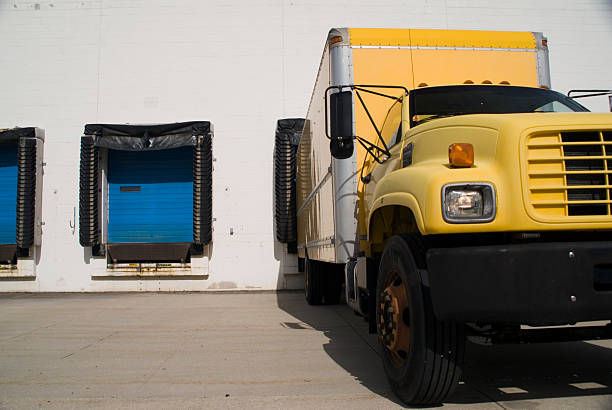 Yellow truck parked at a loading dock with blue doors and black curtains. Bright daylight with shadows on a concrete surface.