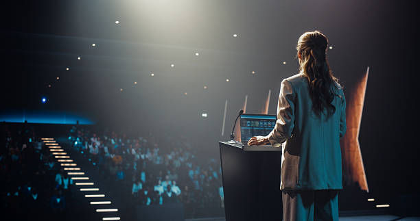 A woman in a suit speaks at a podium in a dimly lit auditorium. Audience members are seated, and a spotlight creates a dramatic mood.