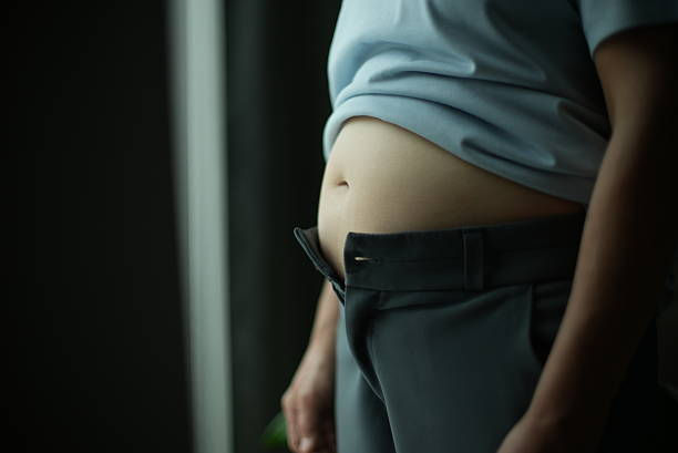 A person stands indoors, wearing unbuttoned gray pants and a light blue shirt, showing a visible belly. The mood is casual.