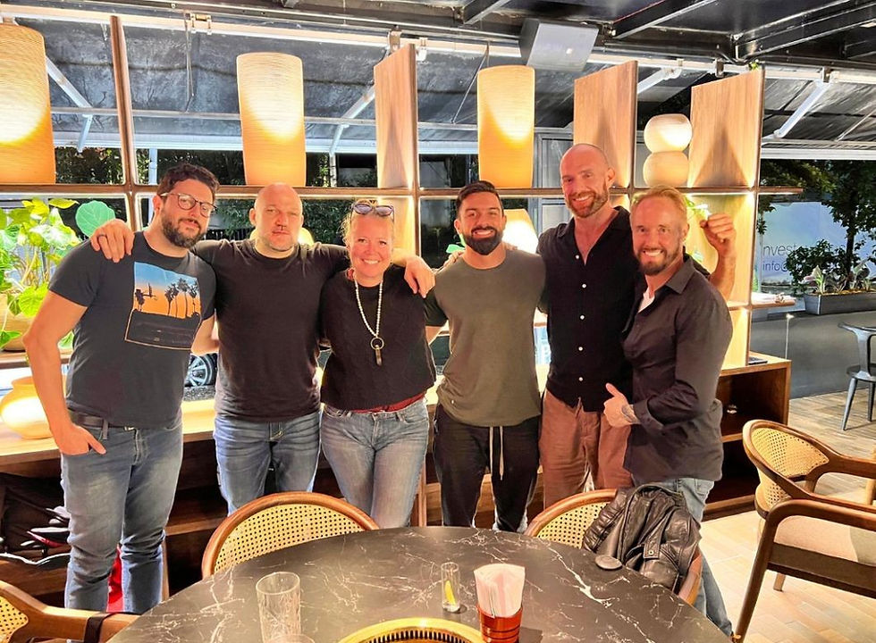 Six people smiling and posing together in a warmly lit restaurant. Background has wooden shelves with lamps and plants. Casual attire.