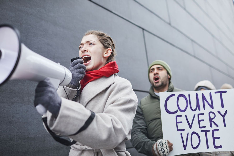Woman shouts into a megaphone at a protest while a man holds a COUNT EVERY VOTE sign beside a gray wall.