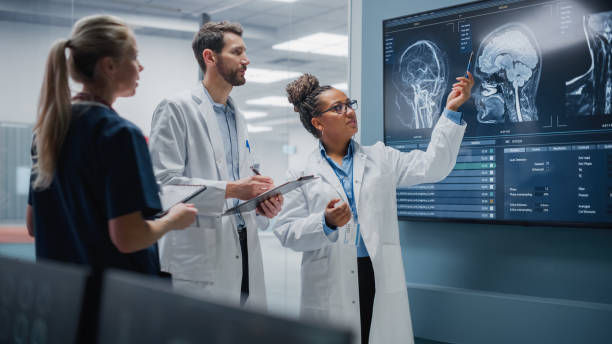 Three doctors in a hospital lab review brain scans on a large monitor, one pointing at the X-ray images, focused and serious