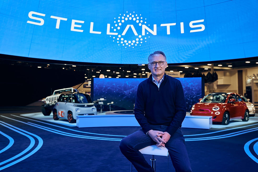 Man seated at Stellantis auto show booth, with small concept cars and bright blue Stellantis logo backdrop behind him.