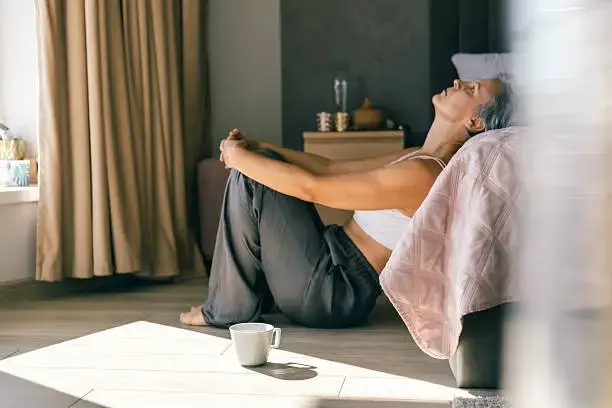Woman sits on a bedroom floor leaning against a bed, knees hugged, with a white mug in sunlight, looking tired and reflective