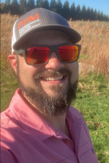 Man in a pink shirt, sunglasses, and hat with "Proactive Risk Management" text smiles outdoors in a sunny field with tall grass.