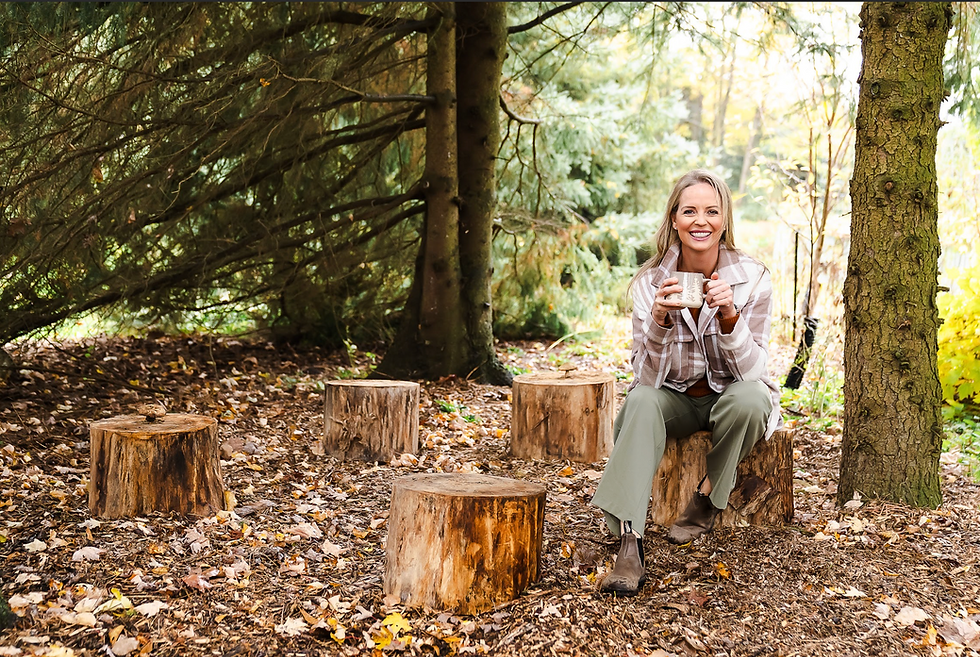 Smiling woman sits on tree stump in forest, holding a mug. Surrounded by trees and fallen leaves, she wears a plaid jacket and green pants.