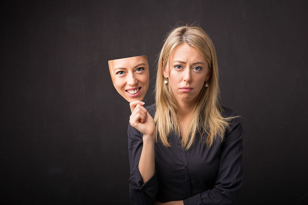A woman in a black shirt holds a smiling mask in front of her; she looks sad. Dark background, conveying a contrasting mood.