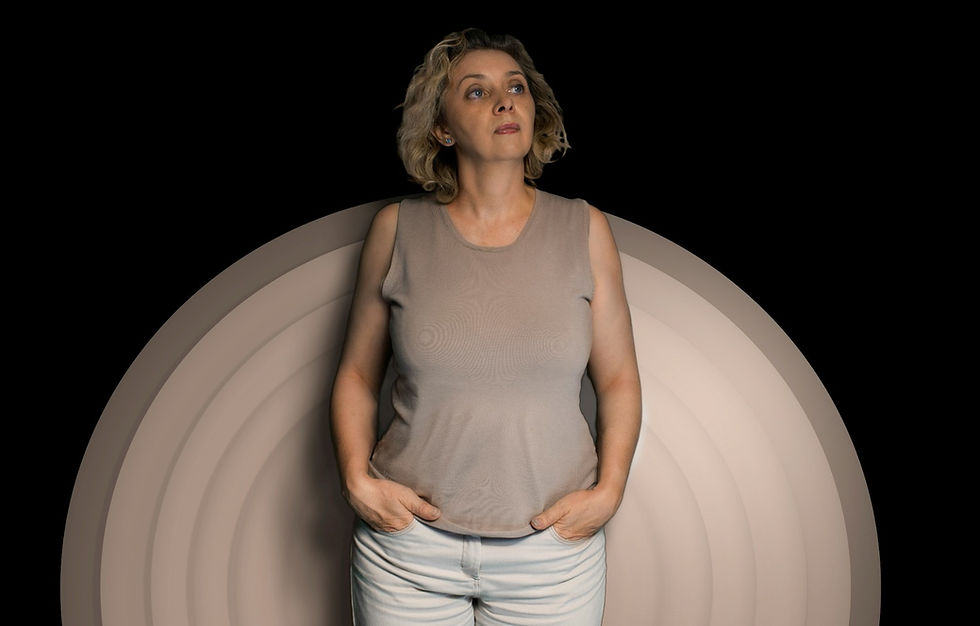 Woman in beige top and light pants stands with hands in pockets against a black background with beige concentric circles, looking thoughtful.