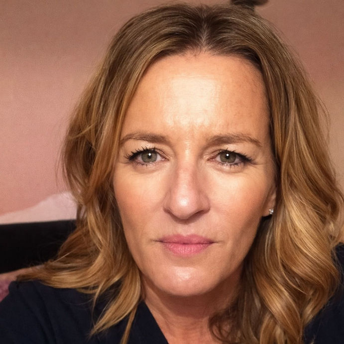 Woman with wavy blonde hair looks directly at the camera. Neutral expression, warm lighting, and a soft pink background.