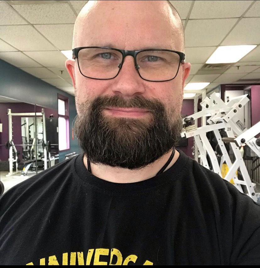 Bald man with a beard wearing glasses and a black "UNIVERSAL" T-shirt smiles in a gym setting with exercise equipment in the background.