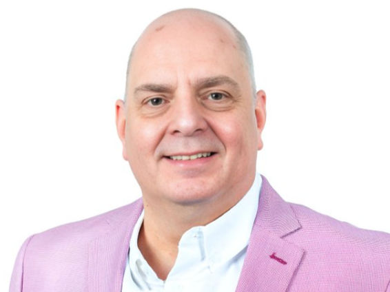 Man smiling, wearing a white shirt and pink blazer, posing against a plain white background. Mood is friendly and approachable.