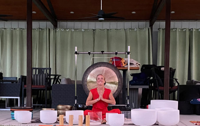 The photo shows a woman dressed in red, sitting cross-legged on the floor with her hands in a prayer position, smiling.
