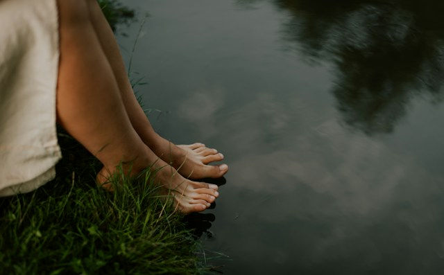 Bare feet rest at the edge of a calm pond beside grass, with soft reflections on the dark water.