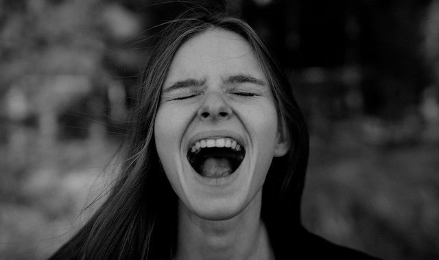 A person with long hair screams with intense emotion. The black-and-white image has a blurred background, enhancing the focus on the expression.