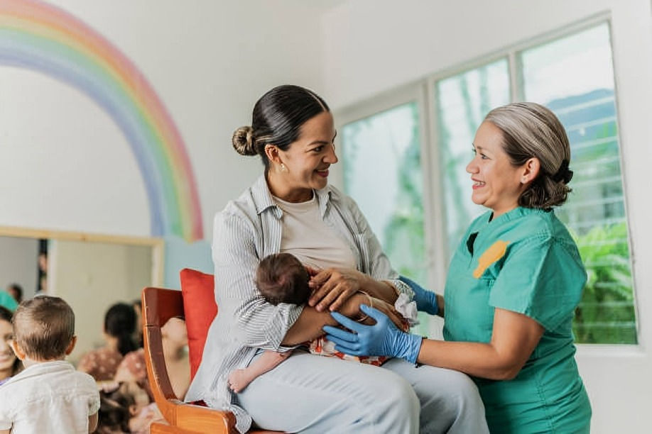 Nurse in teal scrubs comforts a mother holding a baby in a bright clinic with a rainbow mural and large windows.