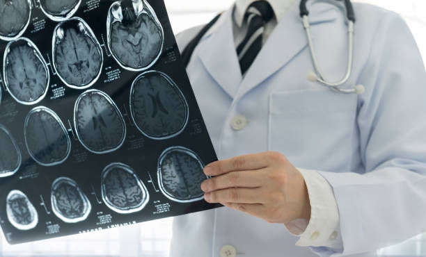 Doctor in white coat and stethoscope examines brain scans on an X-ray film. Background is bright, clinical setting. Focused and professional.