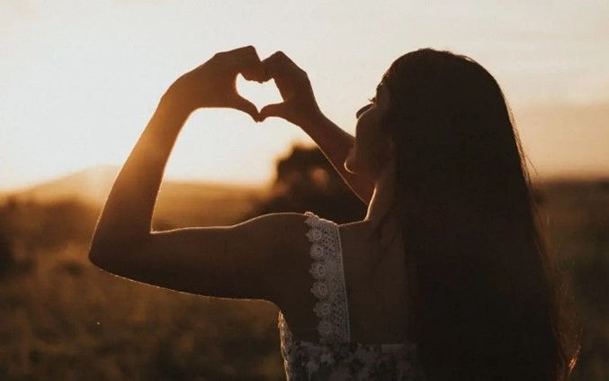 a woman silhouetted against a warm sunset, forming a heart shape with her hands. The sunlight creates a soft glow, adding a sense of peace and positivity to the scene.