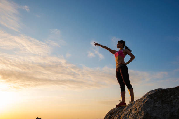 Woman in pink athletic wear stands on a rocky hill at sunrise, pointing into the distance under a clear blue sky.