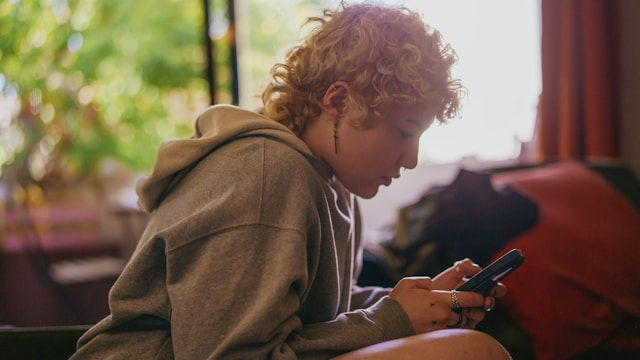 A young person sits indoors, focused intently on their phone, framed by soft natural light from a nearby window.