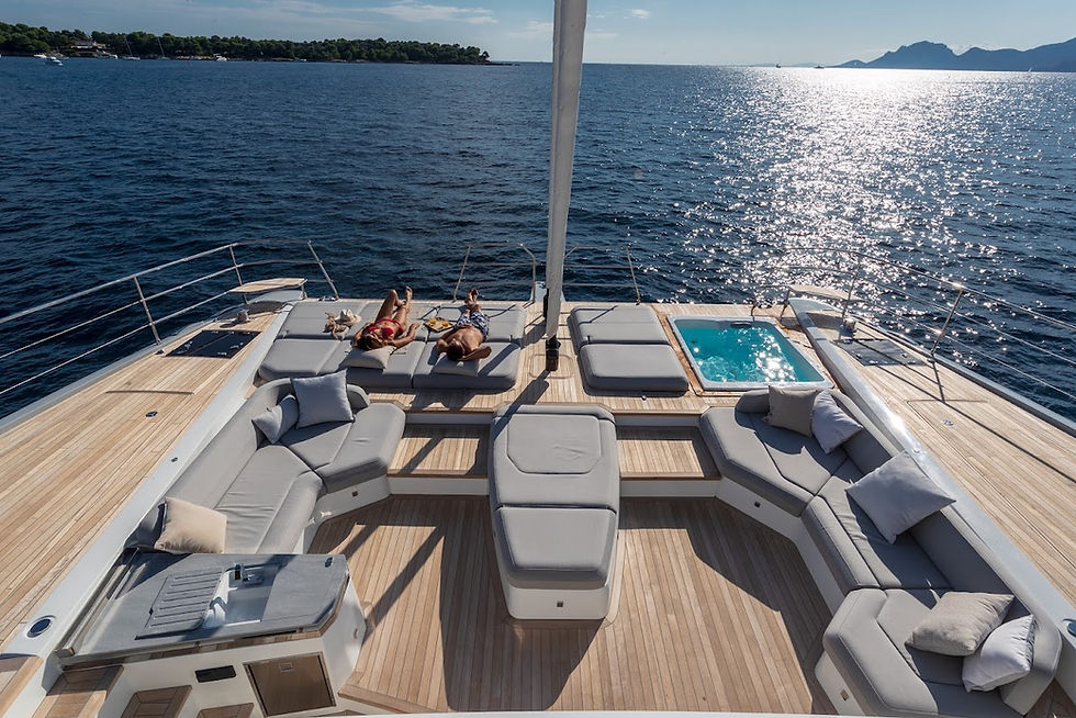 Two people relax on a yacht's sun deck, surrounded by cushions. A small pool is nearby. The ocean and distant hills are in the background.