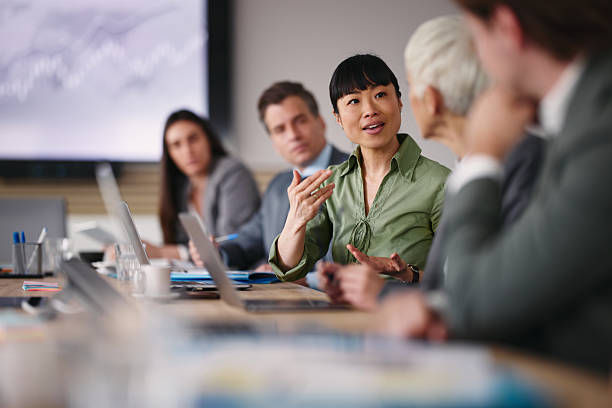 Group of professionals in a meeting room, one woman in a green shirt is speaking. Laptops are on the table. Engaged, focused mood.