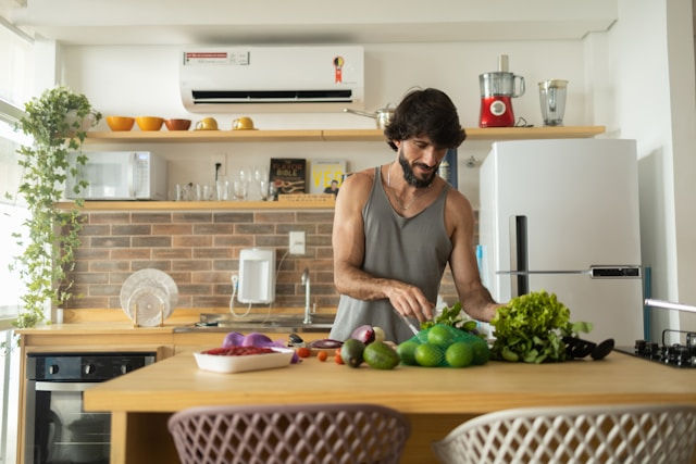 A man with a beard is preparing a healthy meal in his modern kitchen. He's chopping vegetables and has various fruits and vegetables on the counter.