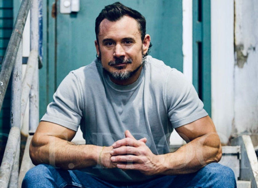 Muscular man in a gray T-shirt sits on outdoor steps with hands clasped, looking at camera, against a teal door.