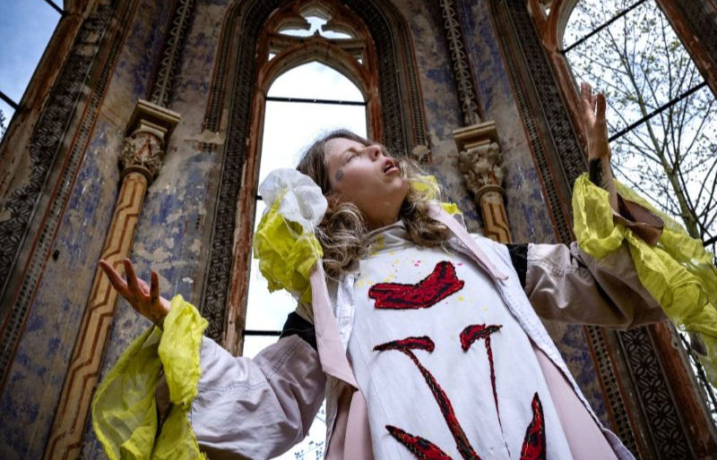 The photo features a person wearing a creatively designed, colorful outfit, standing in an abandoned, ornate space with tall windows and peeling walls.