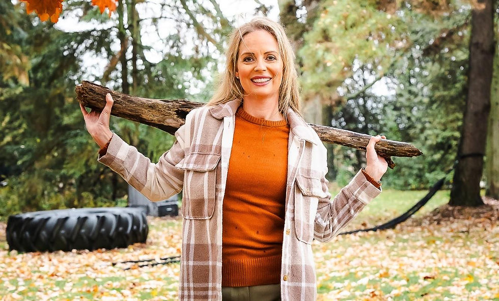 Woman in plaid coat and orange sweater holds a log on shoulders, smiling in a leafy park. Tire in background, autumn mood.