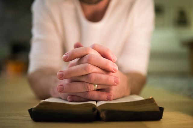 Clasped hands rest over an open book on a table, suggesting quiet prayer in a softly lit room.