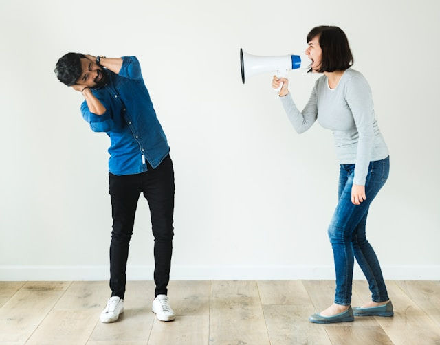 Woman in gray shirt yelling through a megaphone at a man in a blue shirt. He covers his ears, standing on a wooden floor against a white wall.