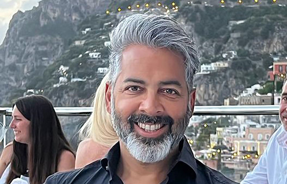Smiling man with gray hair in black shirt in scenic outdoor setting, mountains and buildings in the background. People socialize nearby.