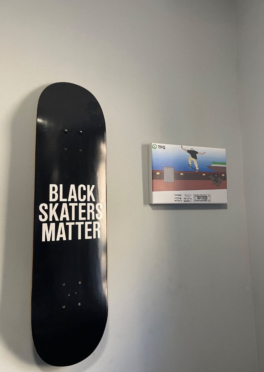 Black skateboard with "Black Skaters Matter" text on a light wall. Next to it, a colorful skateboarding canvas with "TFG" logo.