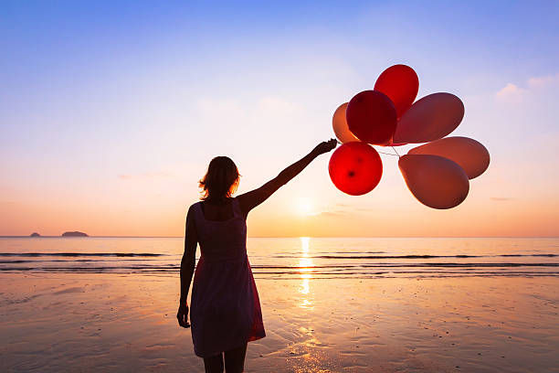 The image shows a silhouette of a woman standing on a beach at sunset, holding red and white balloons.