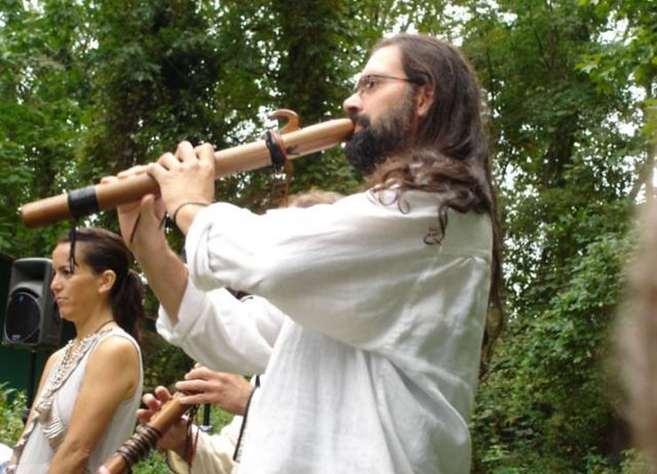 A man in a white shirt plays a wooden flute outdoors, surrounded by greenery. A woman stands nearby, looking attentive.