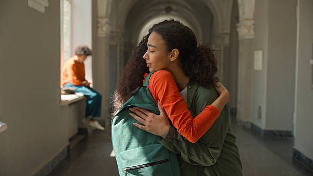 Two young women hug in a sunlit hallway, one wearing a teal backpack and orange sleeves, with a seated person blurred in back.