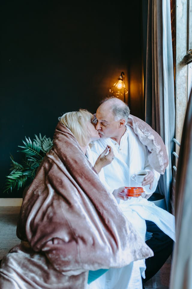 Couple wrapped in a brown blanket shares a kiss by a window, holding a cup and snack. Warm light, cozy mood, plant in background.