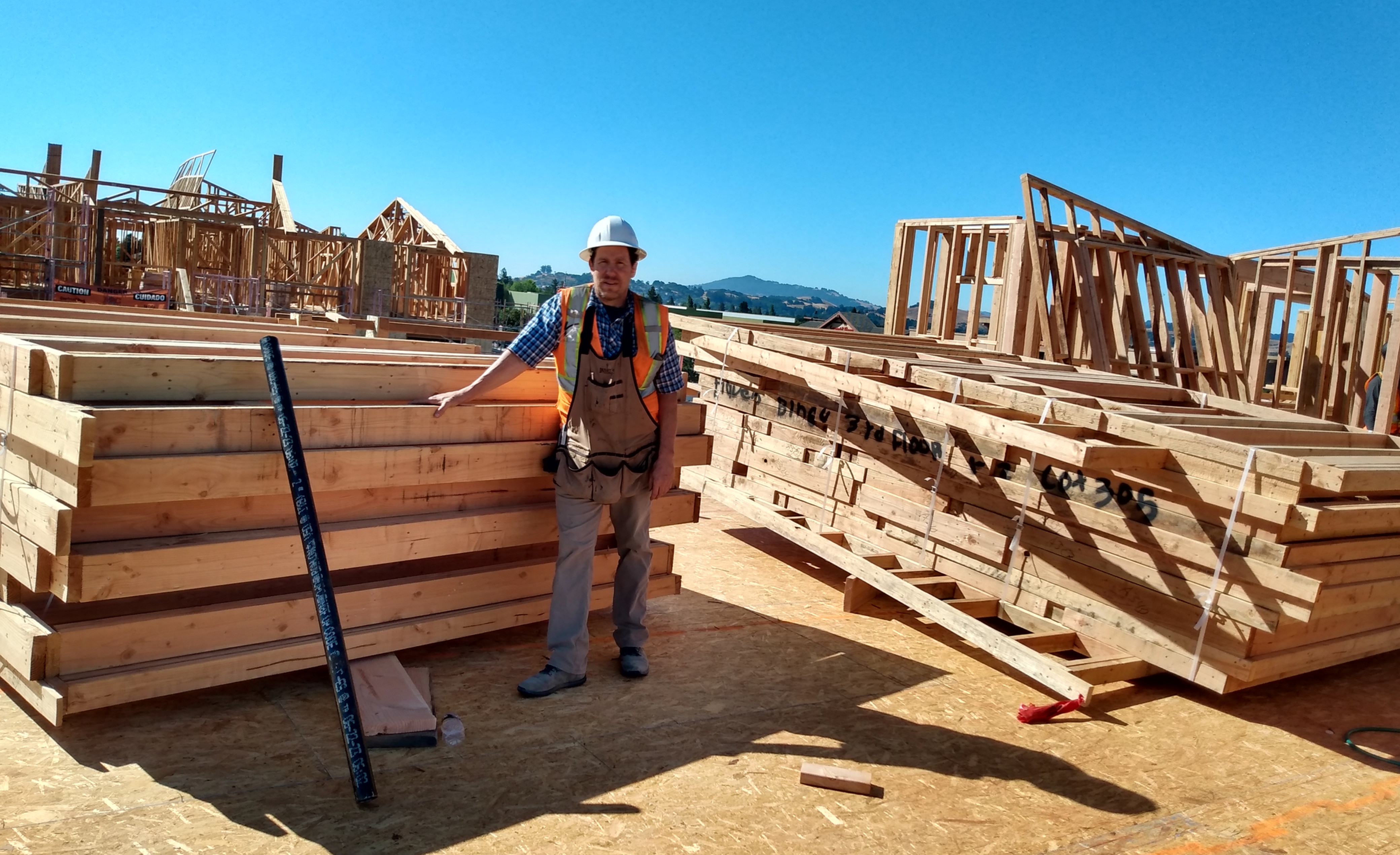 Construction worker in hard hat and safety vest stands among wood house frames at a sunny build site under a clear blue sky.