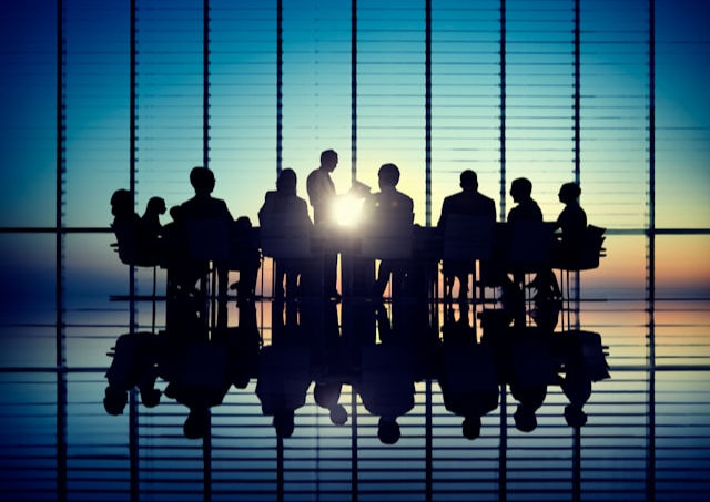 Silhouettes of people in a meeting room with sunset through blinds, creating dramatic reflections on the glossy floor, conveying focus.