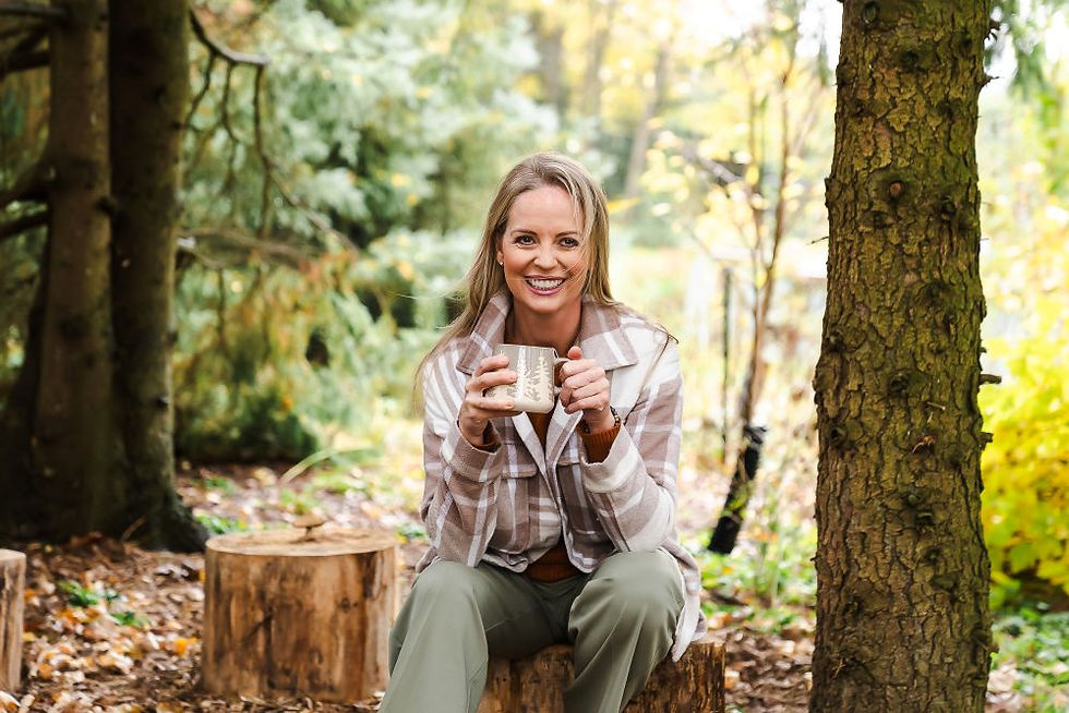 Woman in plaid jacket smiling, holding a mug, sits on a log in a forest setting with autumn leaves and trees in the background.