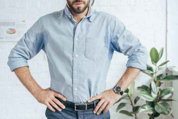 Man in a light blue shirt stands hands on hips in a bright white room beside a leafy plant, looking confident.