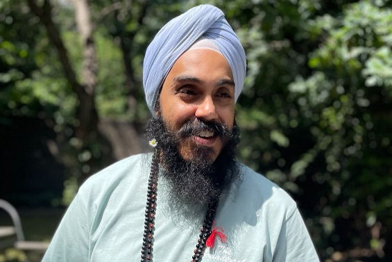 Smiling person with a turban and beard, wearing a striped shirt and beaded necklace, against a plain light background.