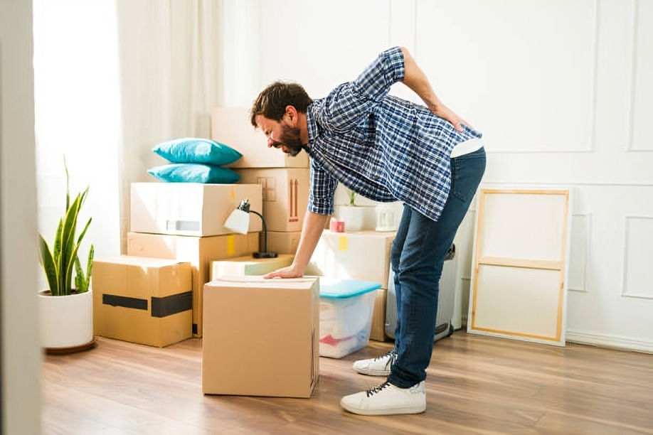 Man in plaid shirt holding his back in discomfort, leaning on a cardboard box in a room filled with moving boxes and plants.