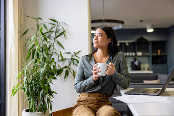 Woman in a green shirt holding a coffee cup, sitting in a modern kitchen with a laptop. Potted plant nearby, looking thoughtful.