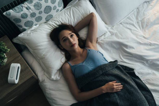 View from top sleepless asian female lying awake on bed is keeping an arm behind head and looking upward while thinking in a dark bedroom